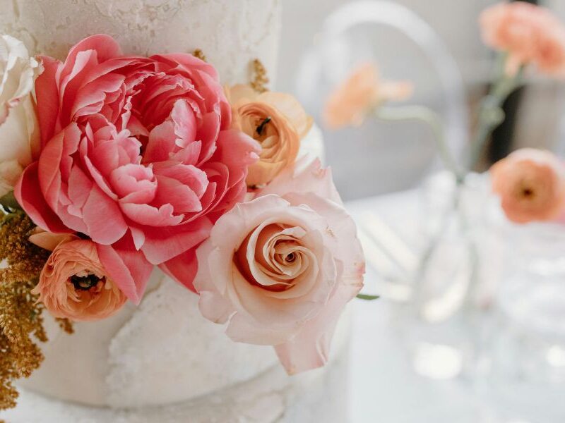 A close-up of a white wedding cake highlights vibrant fresh flowers, including coral peonies, peach ranunculus, and pale pink roses.