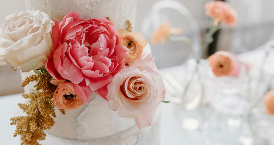 A close-up of a white wedding cake highlights vibrant fresh flowers, including coral peonies, peach ranunculus, and pale pink roses.