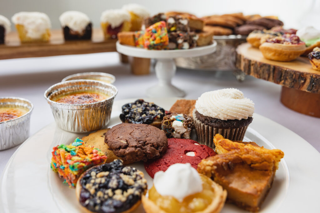 A full dessert bar spread features crème brûlée, red velvet cookies, lemon tarts, cupcakes, and assorted bars and cookies on white platters and rustic wooden stands.