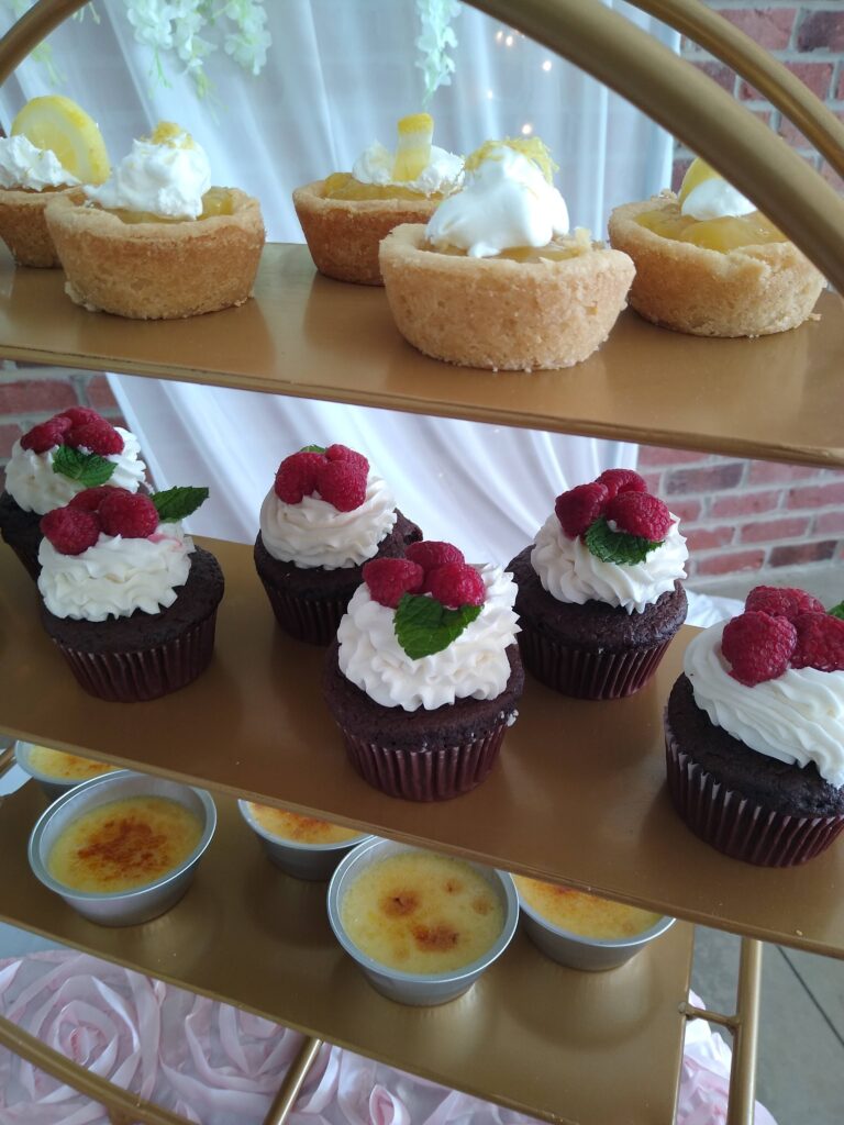 A tiered gold stand displays lemon tarts with whipped cream, chocolate cupcakes topped with raspberries and mint, and cups of crème brûlée on the bottom shelf.