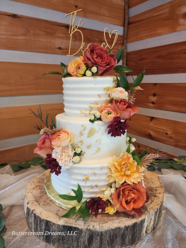 A two-tiered wedding cake from Buttercream Dreams is decorated with textured white frosting, gold accents, and fresh flowers in autumn hues.