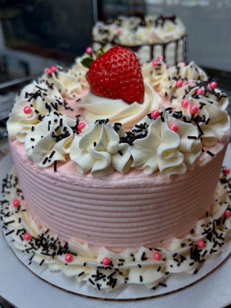 A small pink wedding cake is topped with a fresh strawberry, piped vanilla frosting, pink sugar pearls, and chocolate sprinkles.