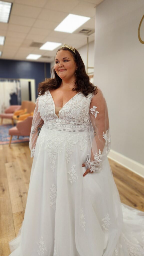 A curvy woman is wearing a white tulle wedding dress with sheer sleeves and an embroidered bodice in a bridal boutique.