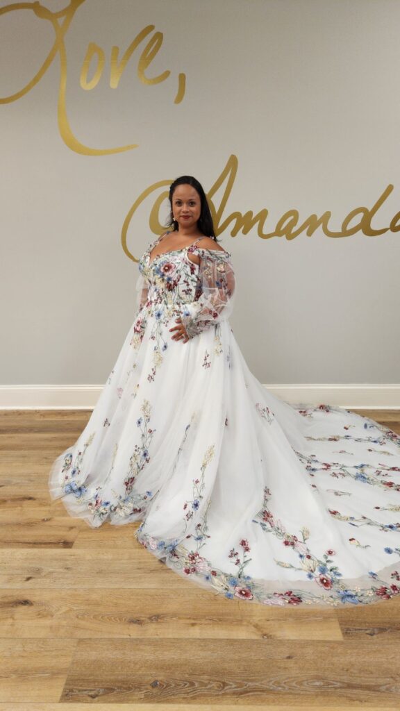 A bride with a curvy figure is wearing a white wedding dress adorned with colorful floral embroidery, standing in front of a wall displaying the words "Love, Amanda".