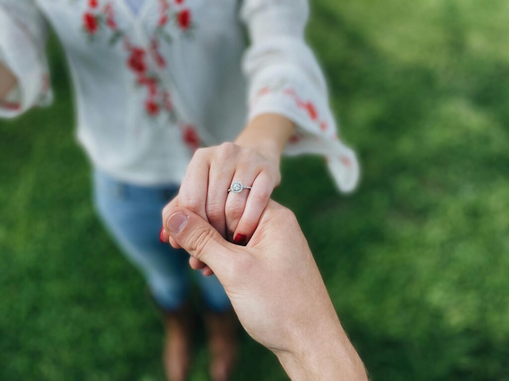 A close-up of two hands holding, with one hand wearing an engagement ring, set against a grassy outdoor background.