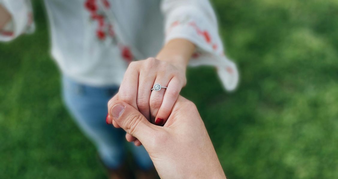 A hand wearing an engagement ring is held by another hand, with a blurred grassy lawn in the background.
