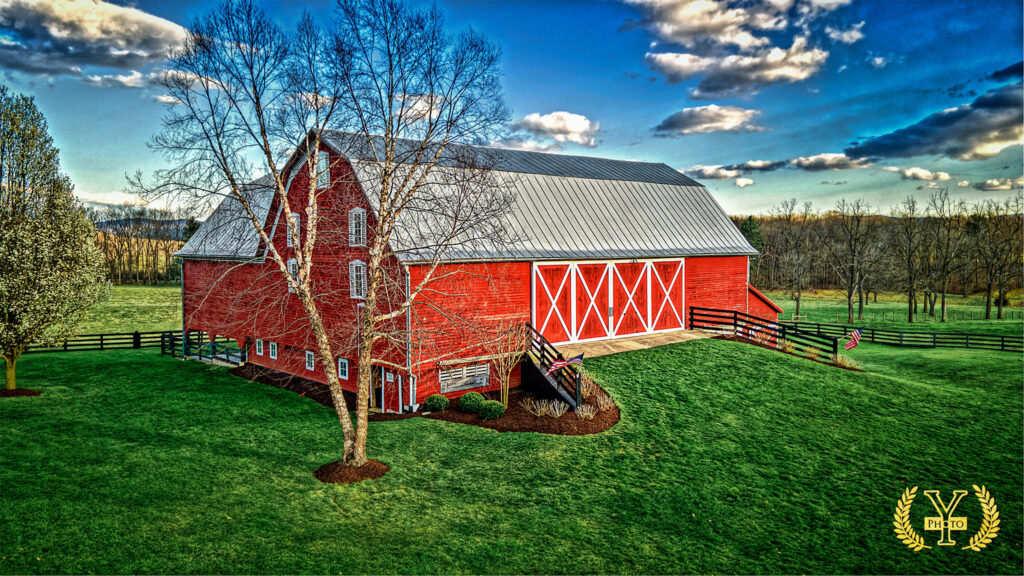 A large red barn with a gray roof sits on a grassy hill, with a bare tree in front and a bright blue sky with clouds overhead.