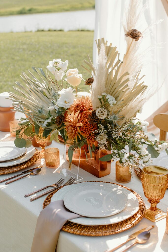 A fall-themed wedding table decorated with a centerpiece of orange, white, and beige flowers, with woven placemats and amber glassware.