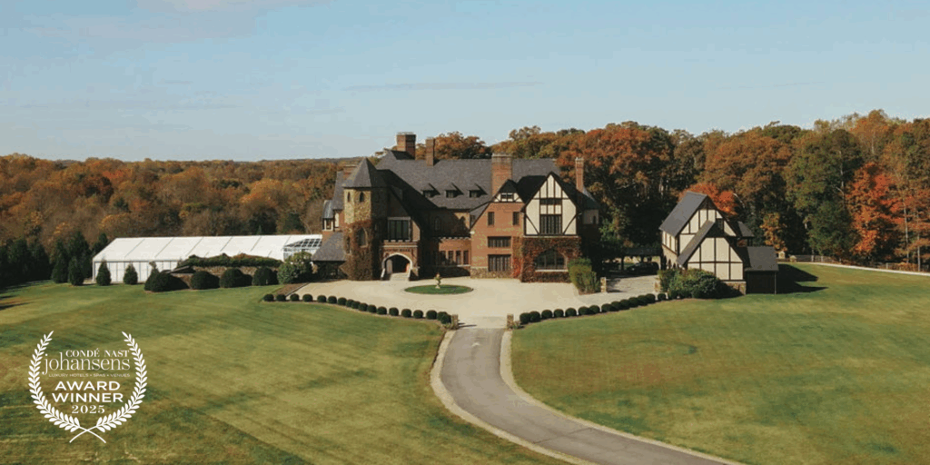 A large brick estate with Tudor-style features is surrounded by expansive green lawns and colorful fall trees in the background.