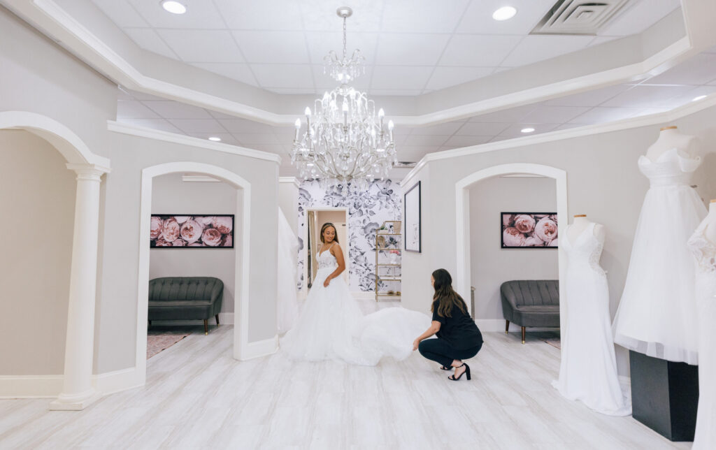 Bride in a white wedding gown standing in a bridal boutique while a stylist adjusts the train, with chandeliers and fitting rooms in the background.