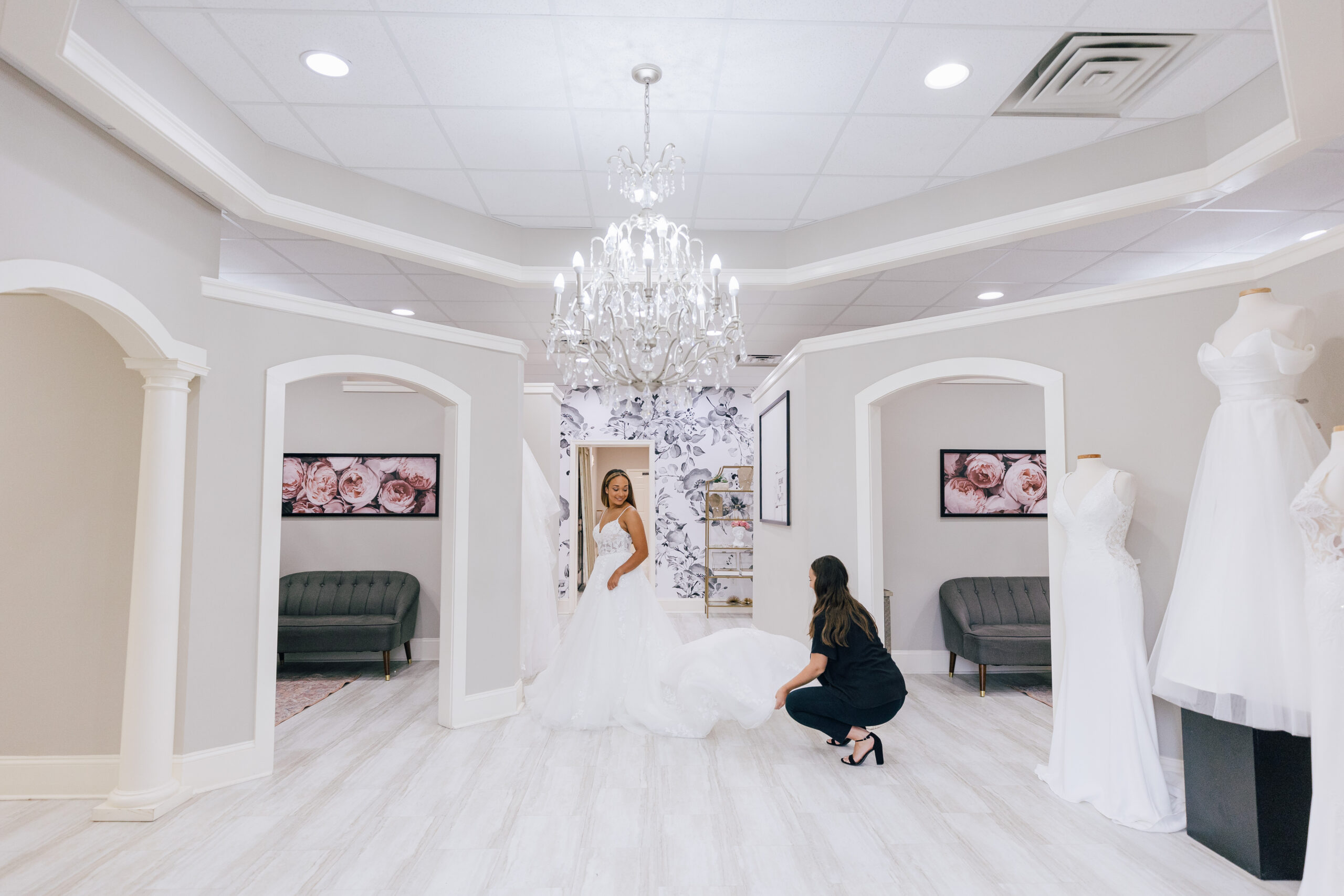 Bride in a white wedding gown standing in a bridal boutique while a stylist adjusts the train, with chandeliers and fitting rooms in the background.