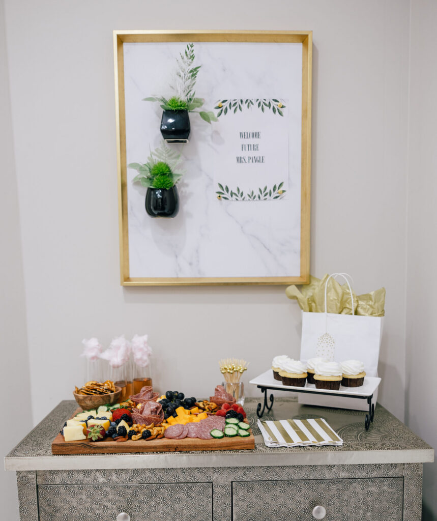 Refreshment table with charcuterie board, cupcakes, gift bag, and welcome sign inside a bridal boutique.