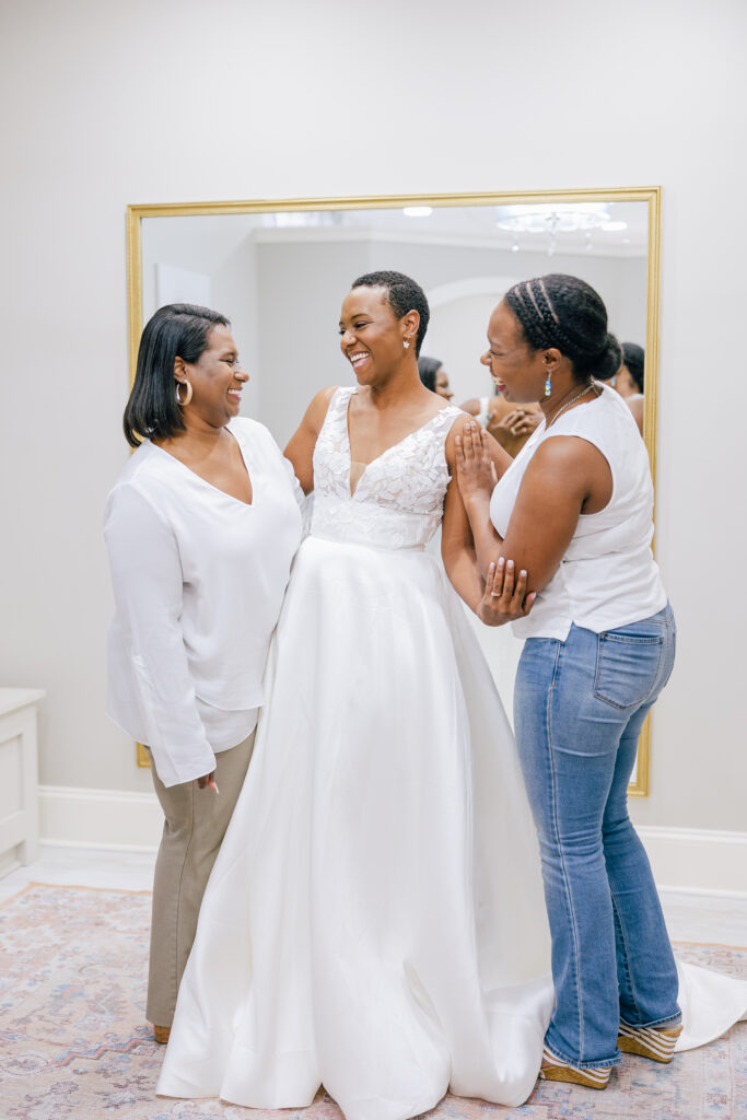 Bride in a sleeveless white wedding gown smiles with two women beside her in a bridal boutique fitting room.