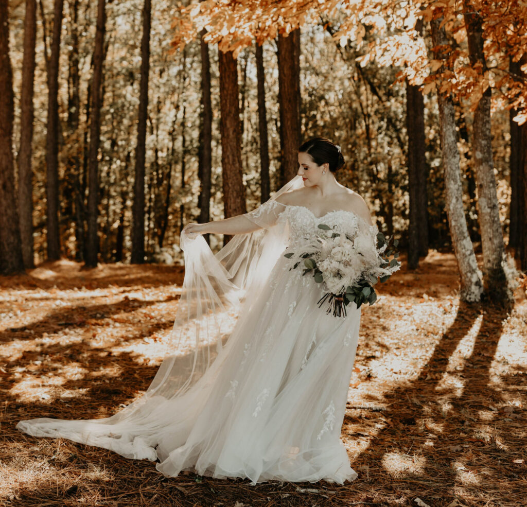 Bride in an off-the-shoulder wedding dress holding a bouquet while standing in a forest with autumn leaves.