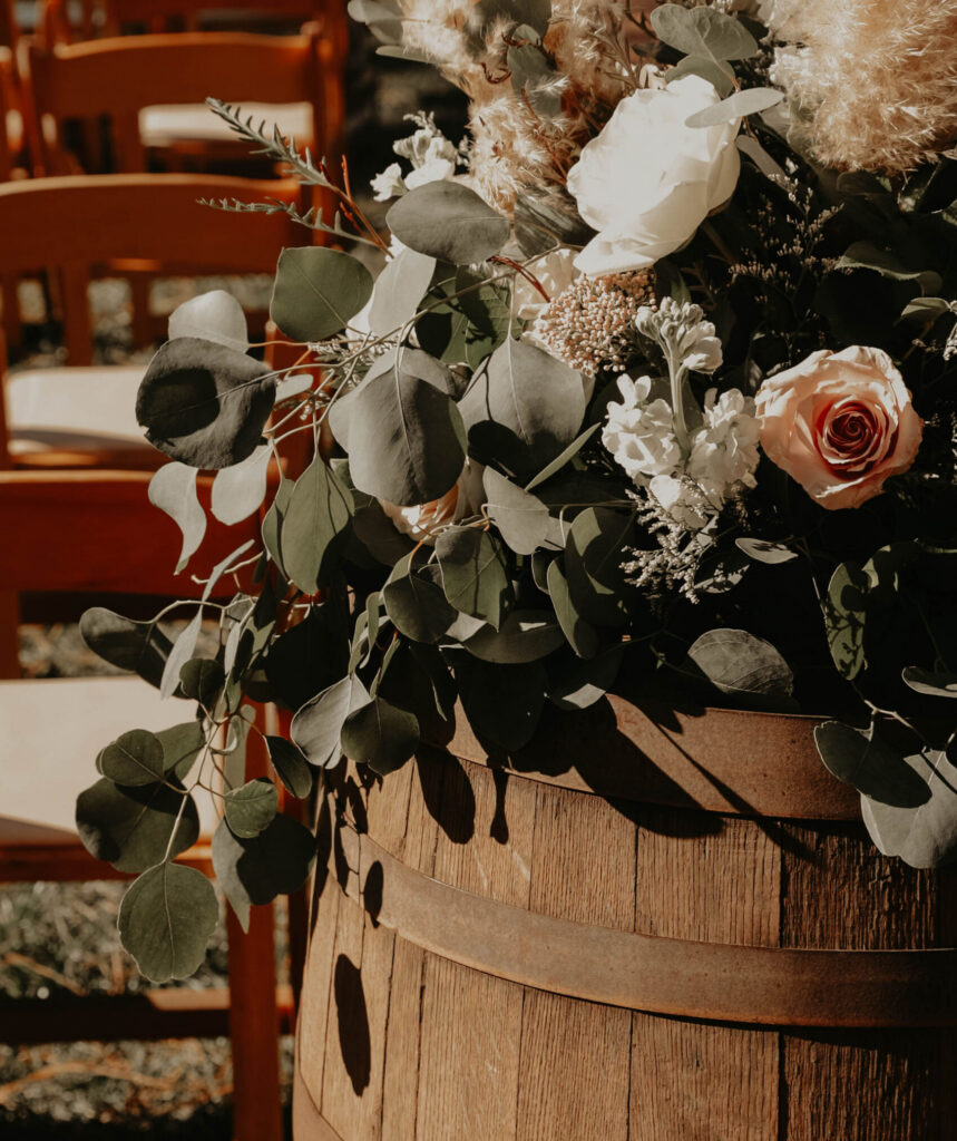 Rustic floral arrangement with pampas grass, roses, and greenery displayed in a wooden barrel beside ceremony chairs.