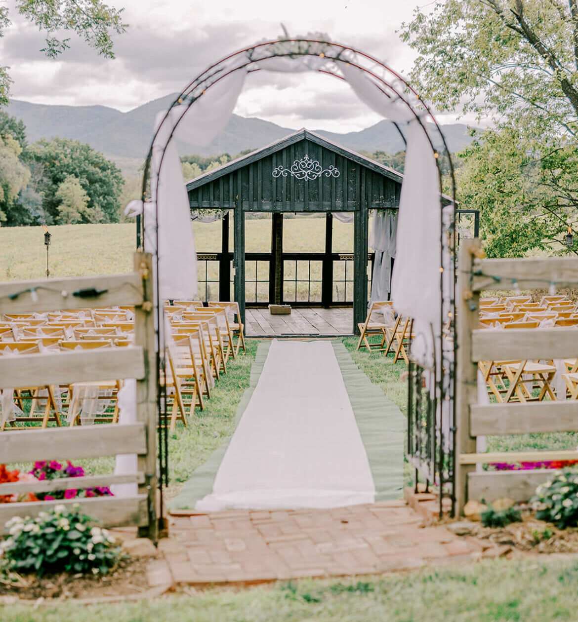 Outdoor wedding ceremony aisle leading through a draped arch toward a dark barn with mountains in the background.