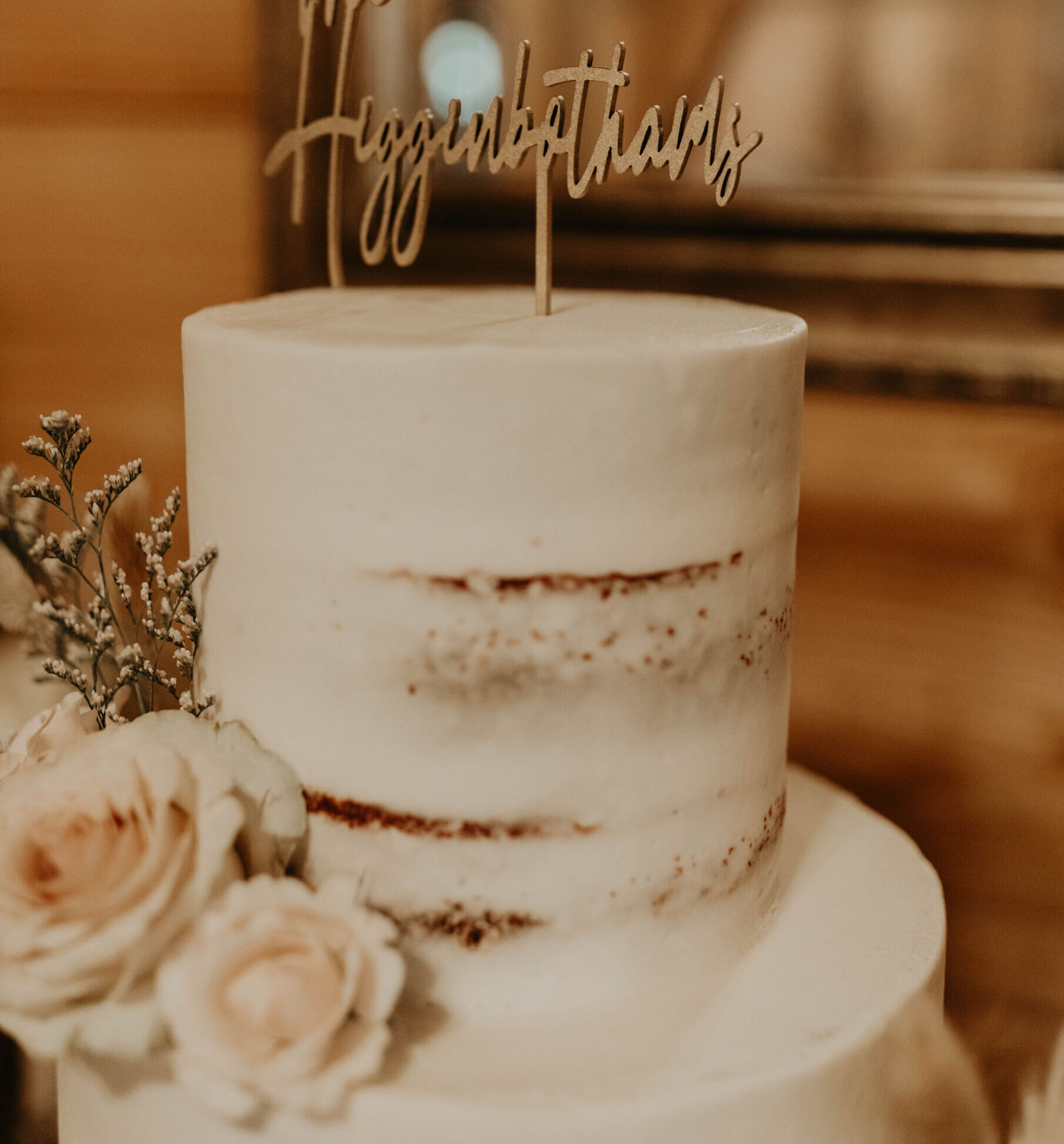 White wedding cake with floral accents and a wooden “The Higginbothams” cake topper on display indoors.