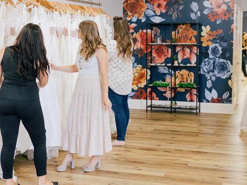 Bridal boutique interior with women shopping for gowns, rows of dresses, floral accent wall, and seating area.