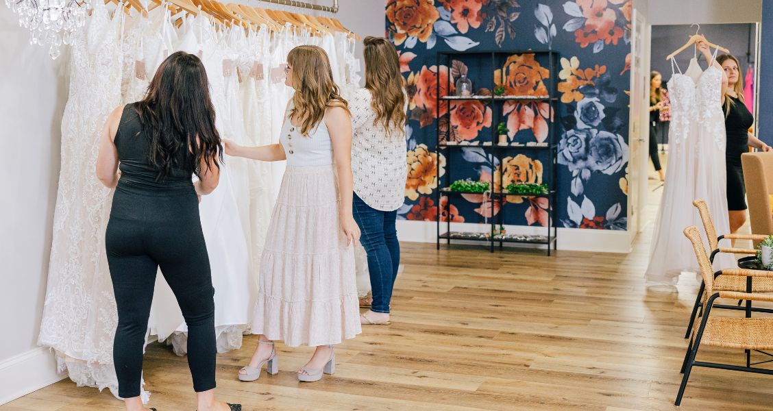 Bridal boutique interior with women shopping for gowns, rows of dresses, floral accent wall, and seating area.