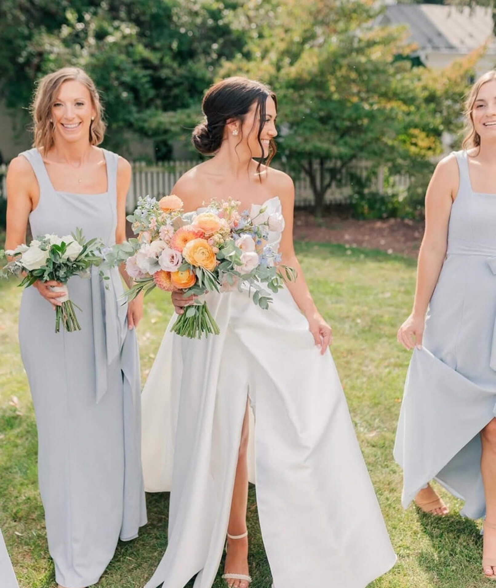 A bride in a satin ballgown with a side slit walks alongside her bridesmaids dressed in light blue gowns, all holding pastel floral bouquets in an outdoor garden setting.