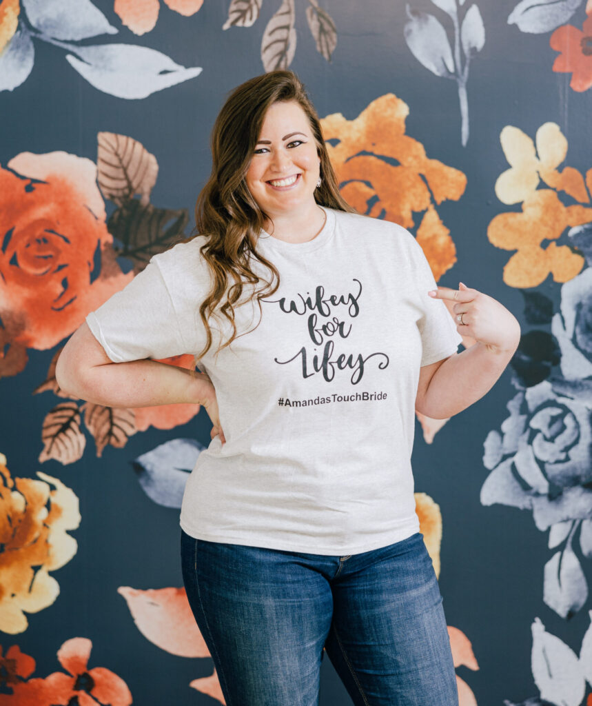 Smiling woman wearing a graphic T-shirt and jeans stands in front of a dark floral mural wall.