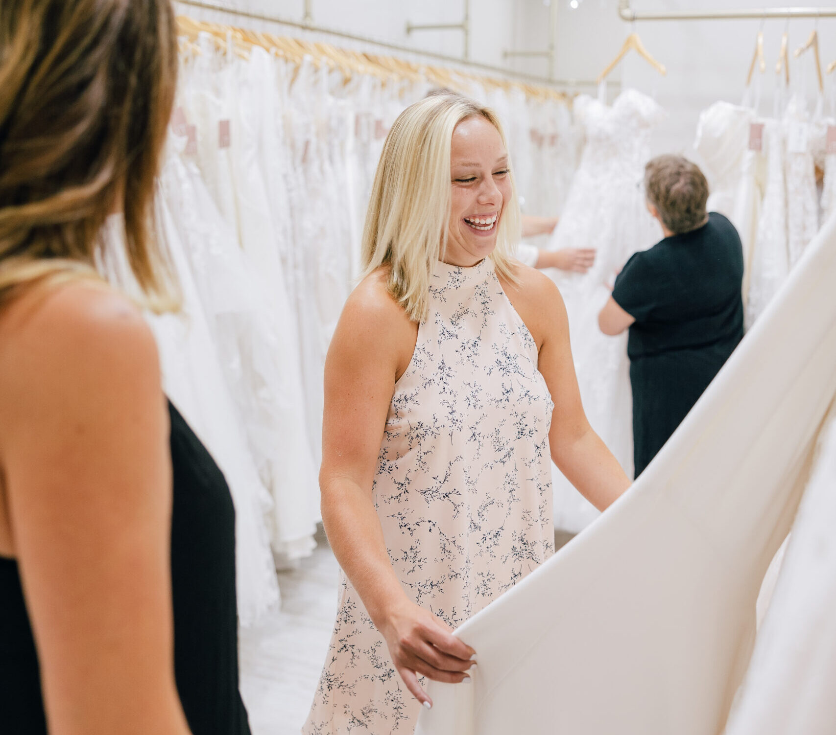 Bride-to-be smiling while holding a white wedding gown during a shopping appointment in a bridal boutique.