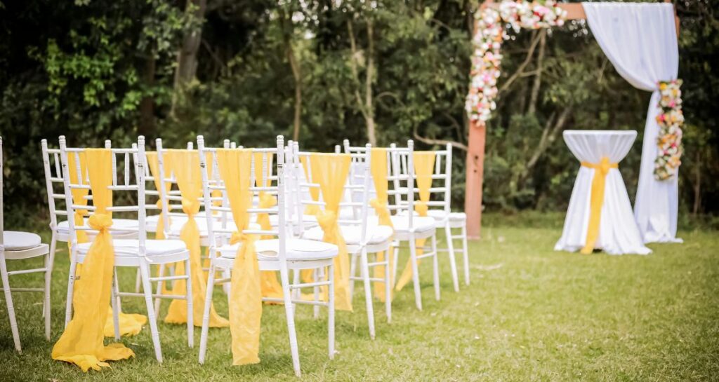 White ceremony chairs decorated with yellow fabric arranged outdoors in front of a floral wedding arch surrounded by trees.