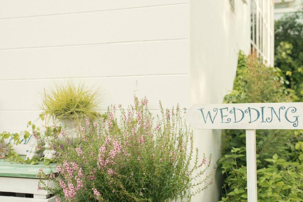 Wooden sign reading “Wedding” beside pink flowers and greenery outside a white garden venue building.