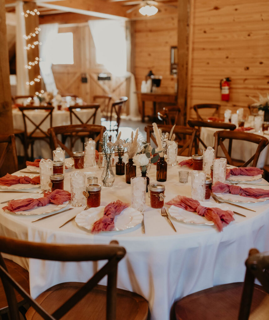 Rustic wedding reception table with floral centerpieces, candles, and pink napkins inside a wooden barn venue.