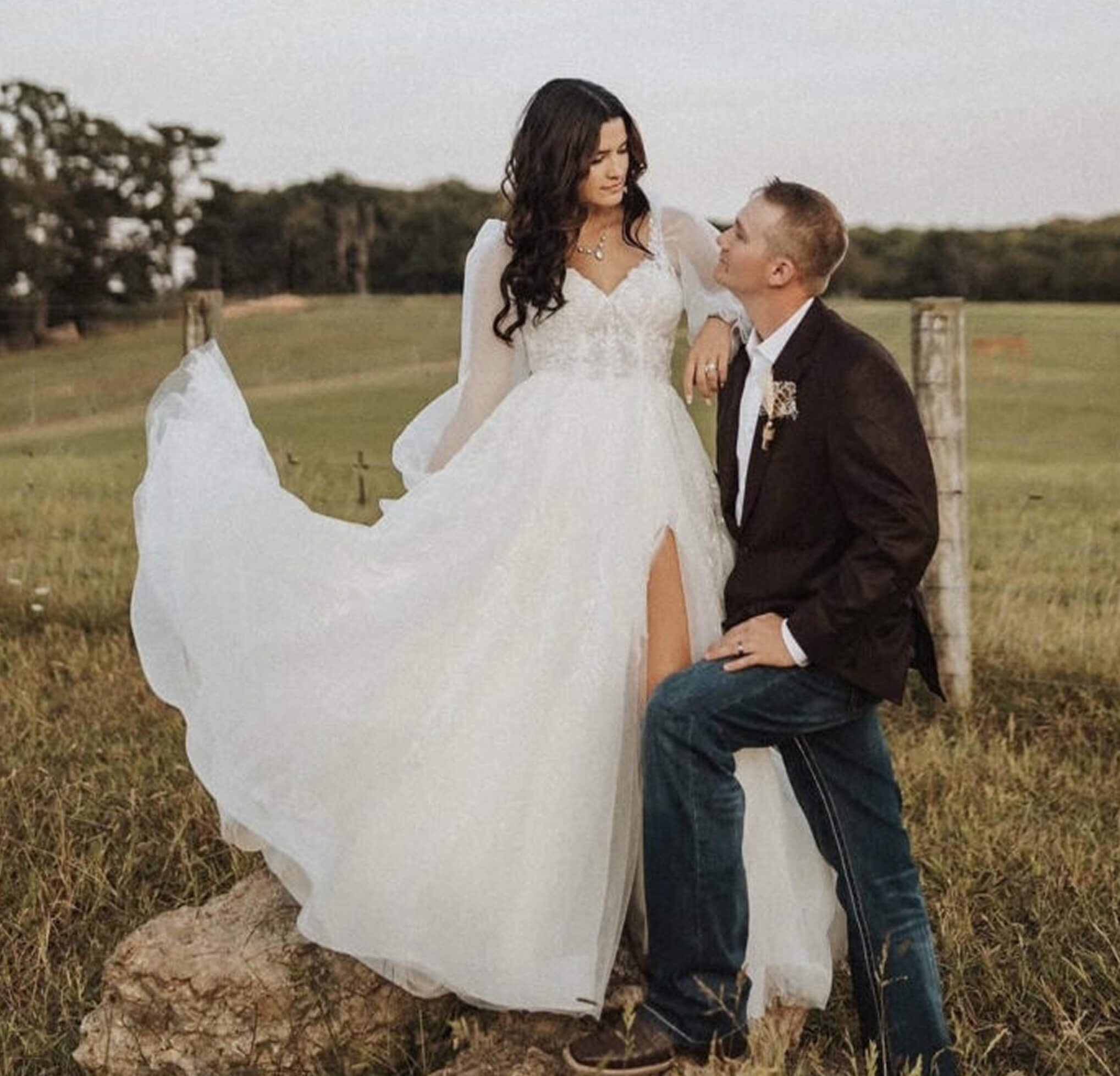 A bride in a long sleeve white gown with a thigh-high slit stands beside her groom in a country field at sunset.