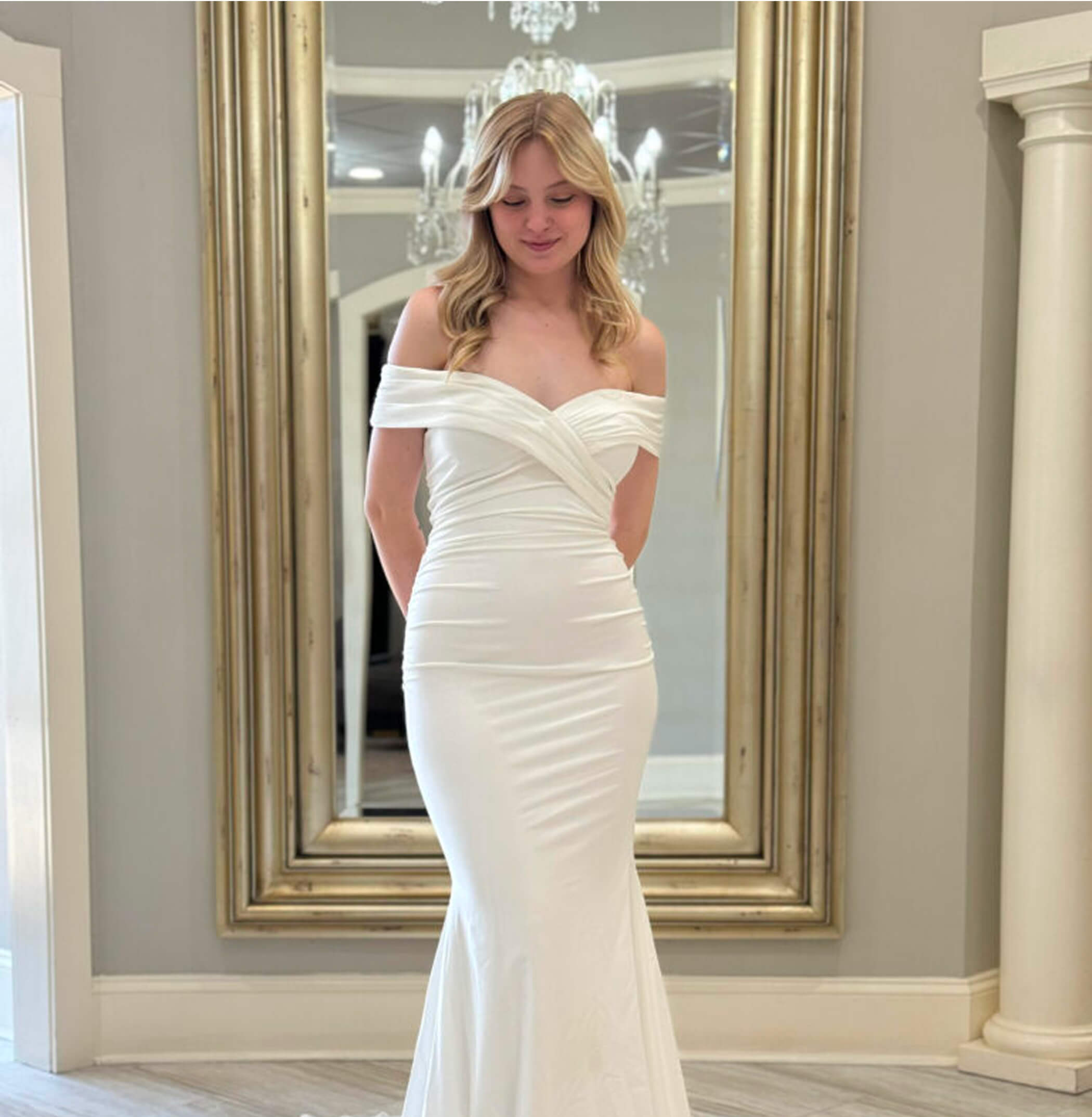 A bride in a sleek, off-the-shoulder fitted gown with a slight flare at the hem, standing in front of a gold mirror and chandelier display.
