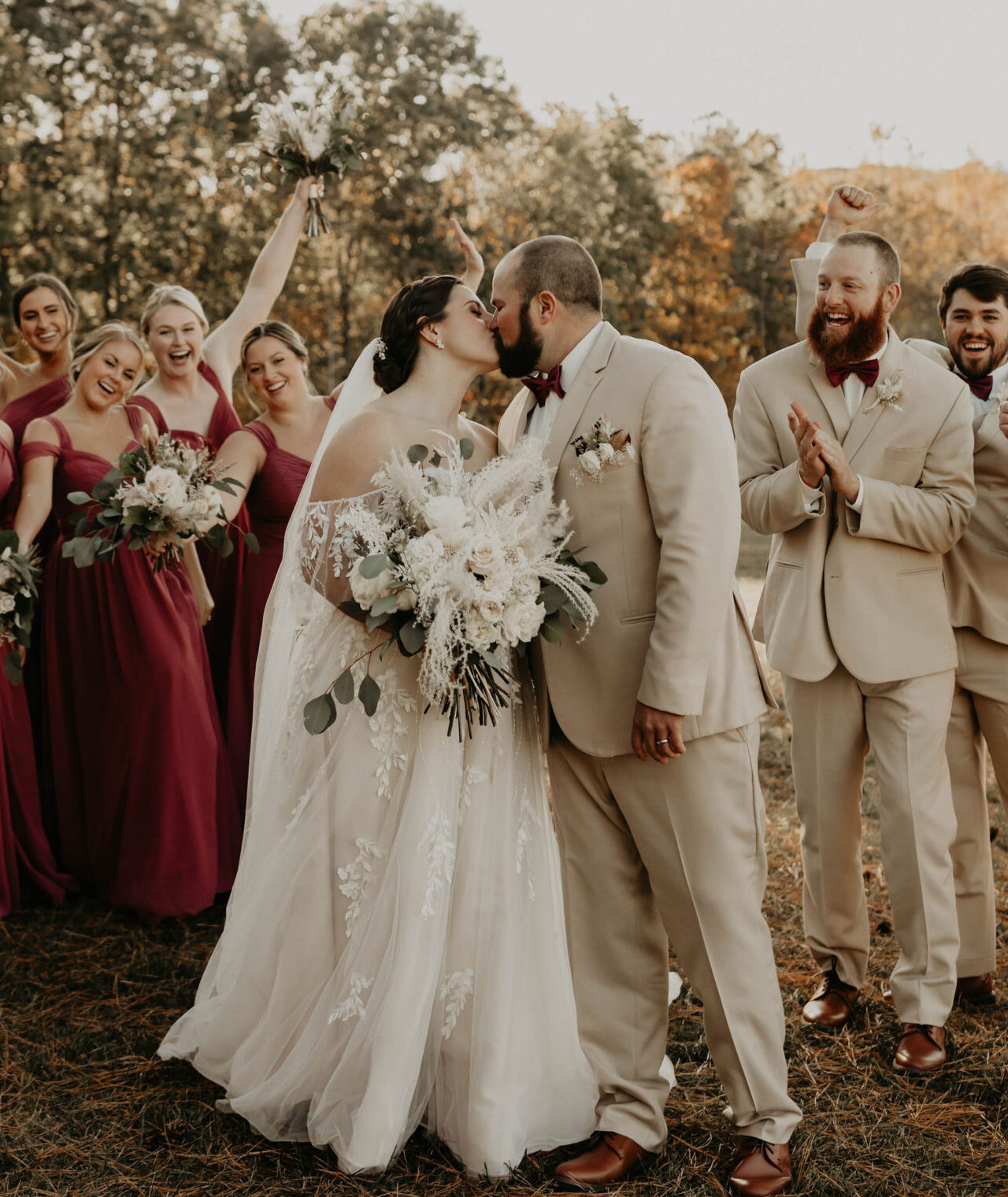 Bride and groom kissing outdoors while bridesmaids in burgundy dresses and groomsmen in tan suits celebrate around them.