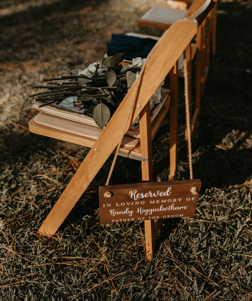 Reserved memorial sign hanging from a wooden ceremony chair beside bouquets during an outdoor wedding ceremony.