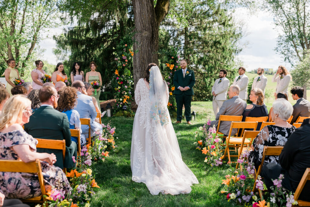 Outdoor wedding ceremony with guests seated on both sides of a flower-lined aisle as a bride walks toward the altar.