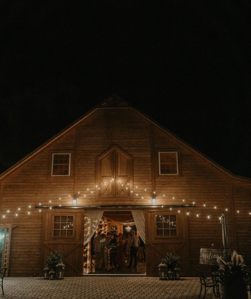Rustic wooden barn wedding venue lit with string lights at night with guests gathered inside the entrance.