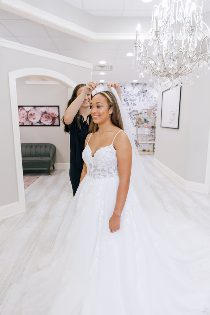 Stylist places a veil on a smiling bride wearing a lace bodice wedding gown beneath a crystal chandelier.