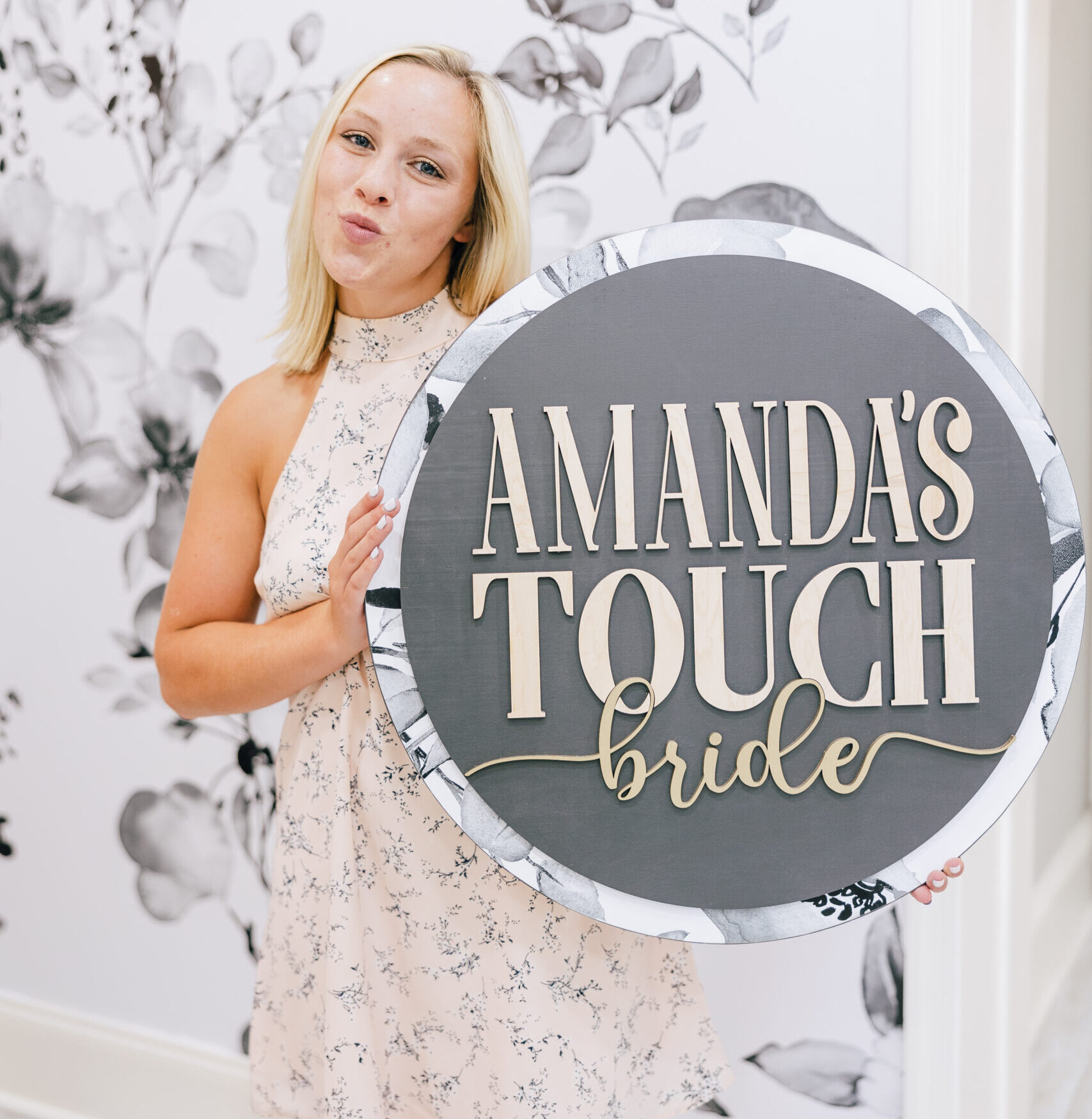 Woman posing with a round boutique sign in front of a black and white floral mural wall.