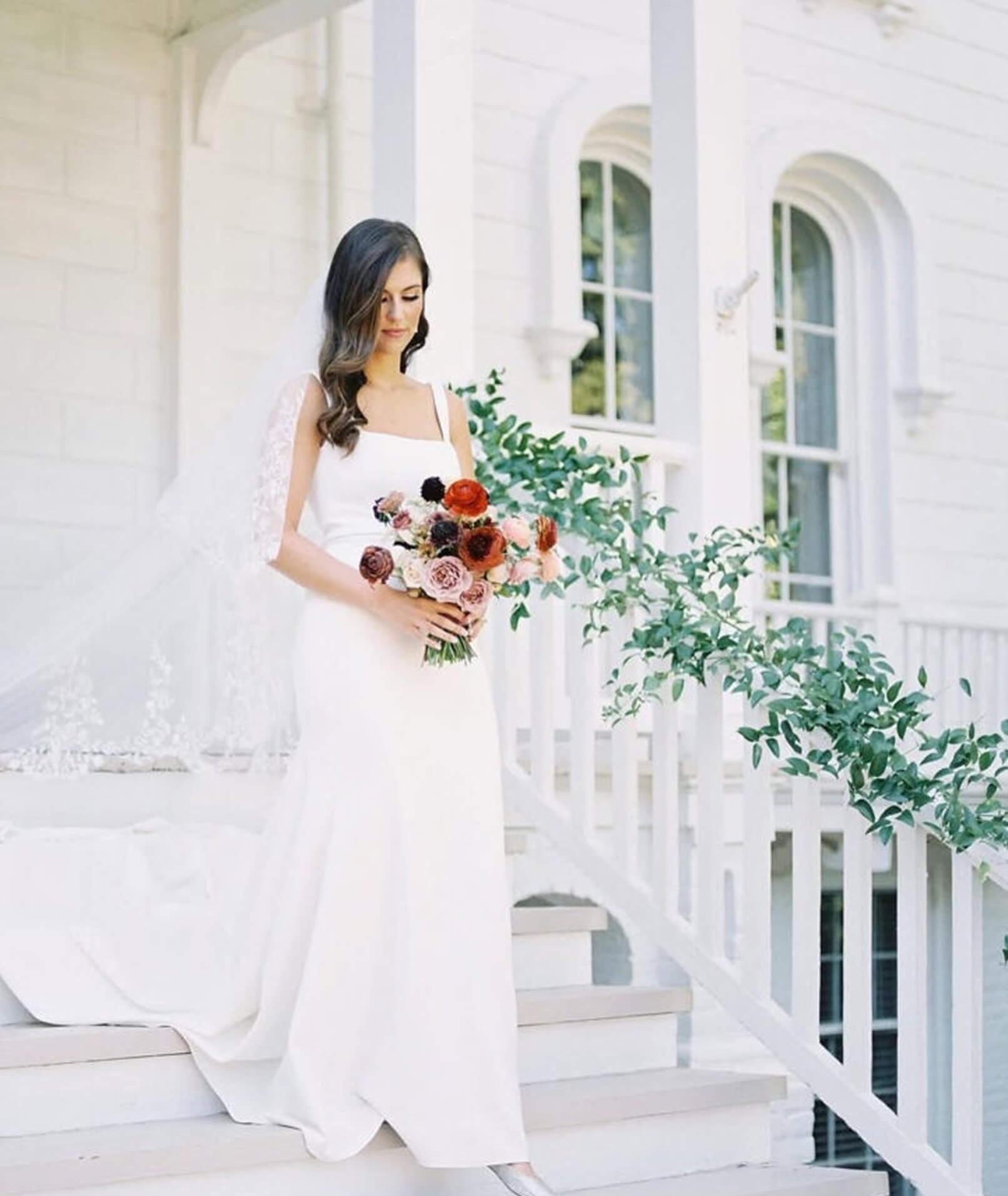 A bride in a minimalist square-neck wedding dress descends white mansion steps, holding a bouquet of red, pink, and plum flowers.