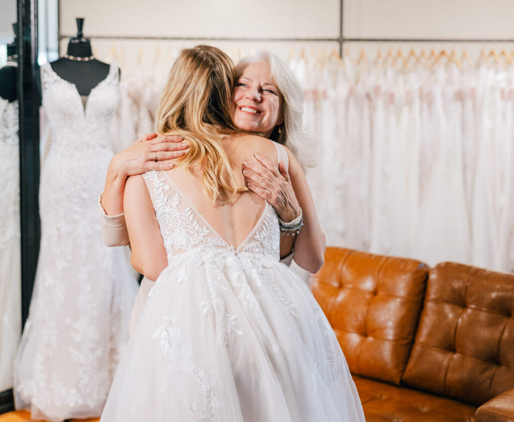 Bride in a lace wedding gown hugging an older woman inside a bridal boutique showroom.