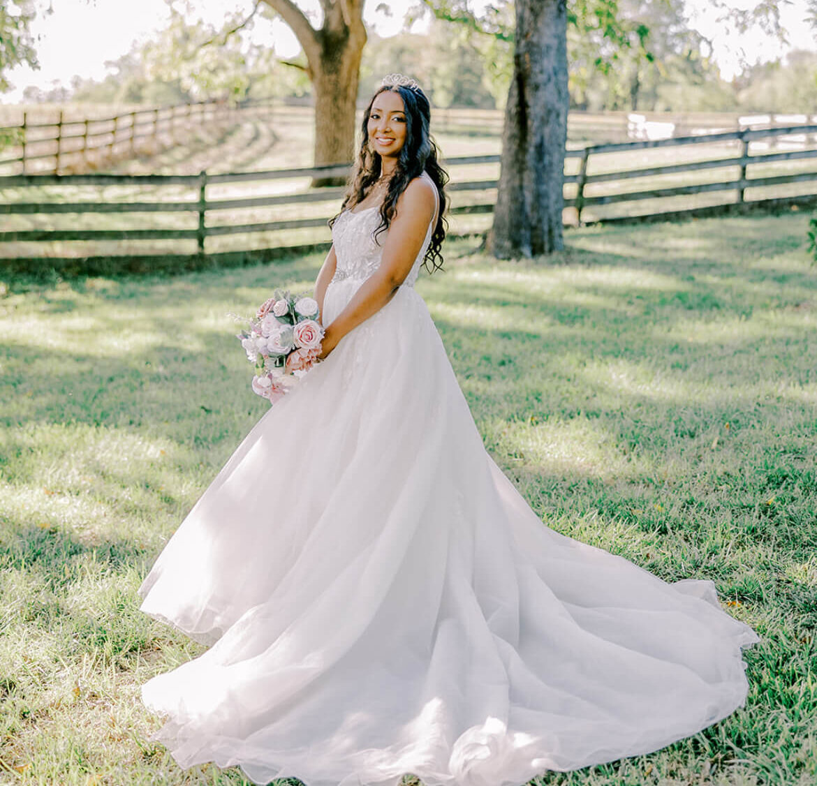 Bride in a flowing wedding gown holding a bouquet while standing beneath large trees in an open field.