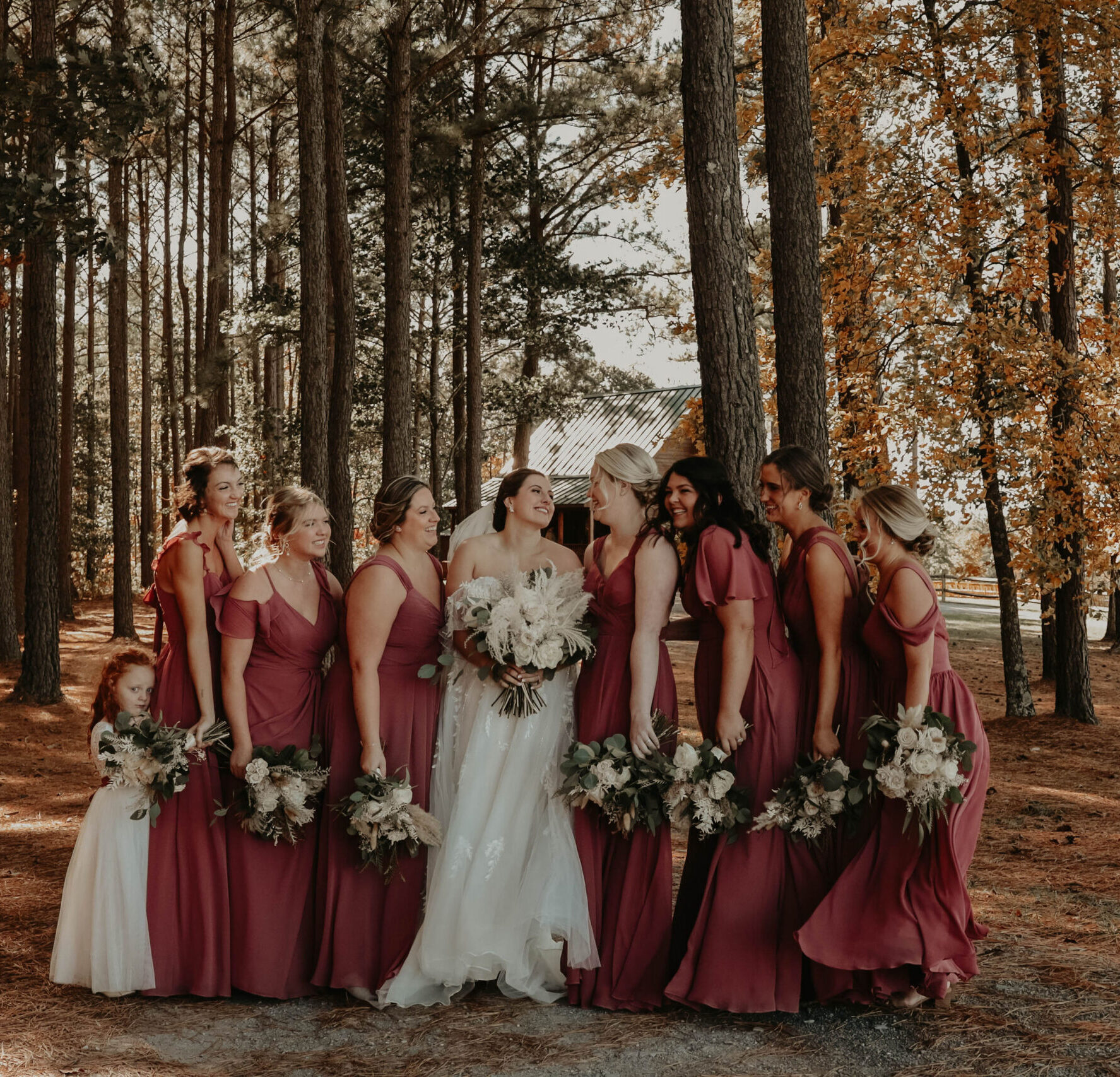 Bride surrounded by bridesmaids in matching burgundy dresses while posing in a forest setting with autumn foliage.