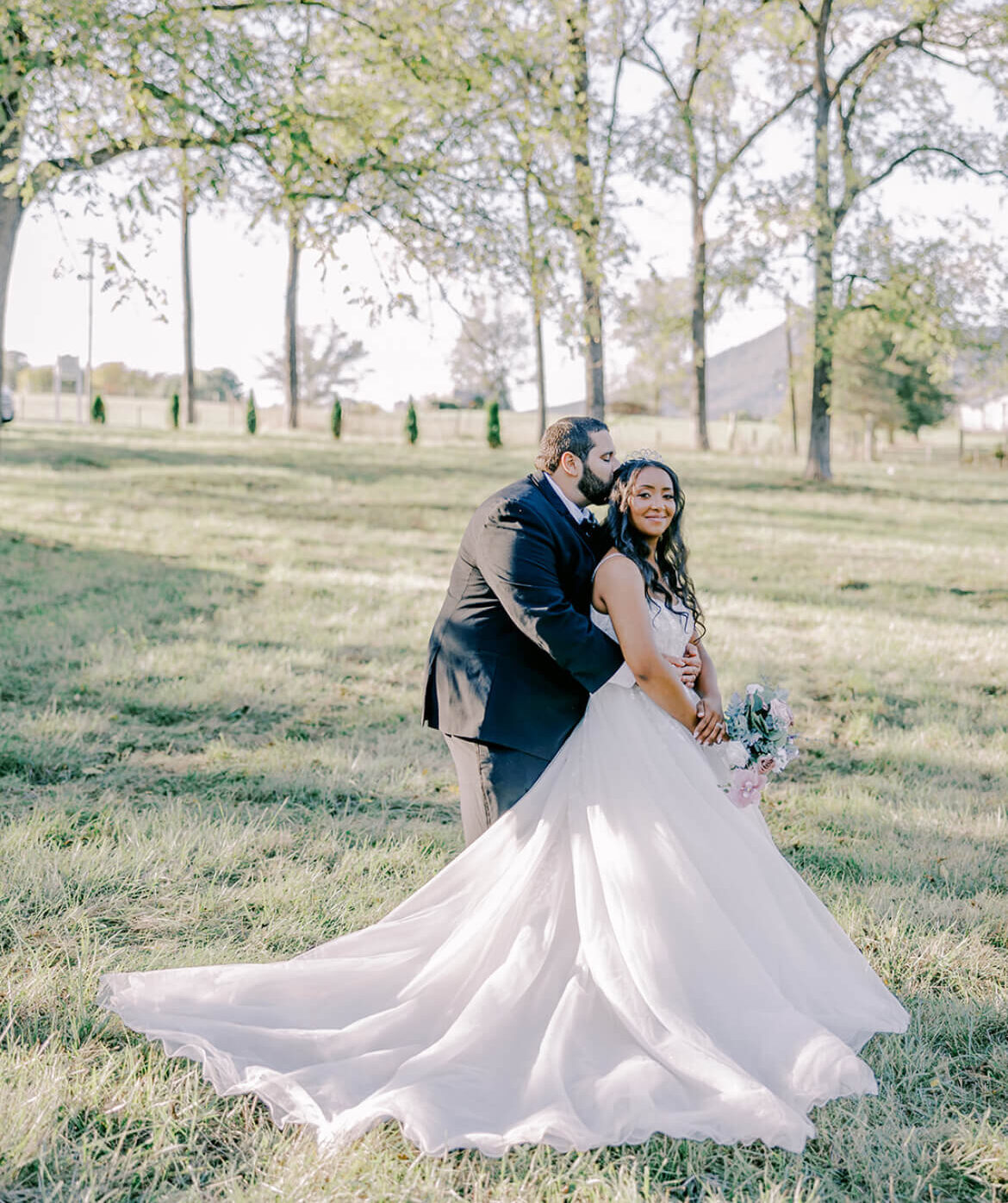 Groom embracing the bride from behind while standing together in a grassy field with trees in the background.