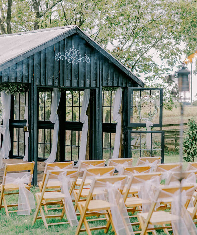 Outdoor wedding ceremony setup with wooden folding chairs facing a black chapel structure decorated with white drapery.