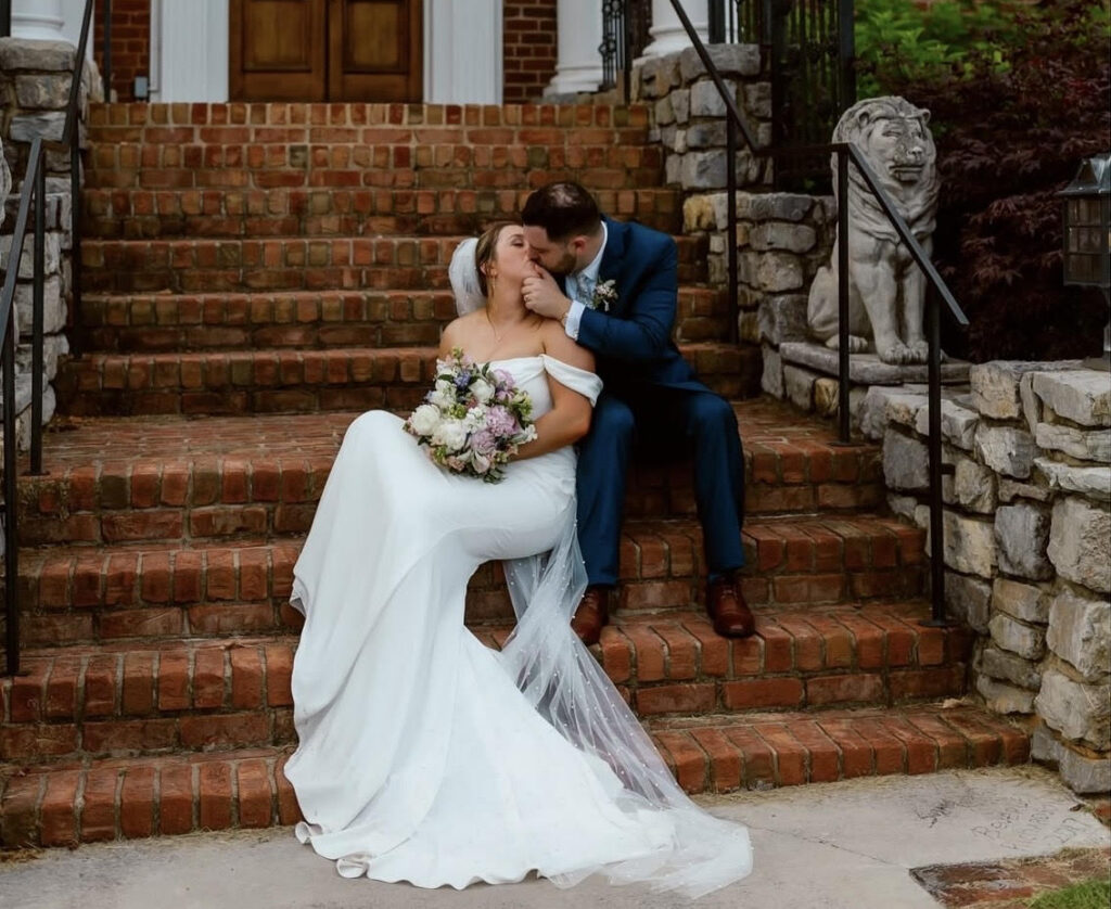 Bride and groom sitting together on brick steps outside a large brick home while sharing a kiss.