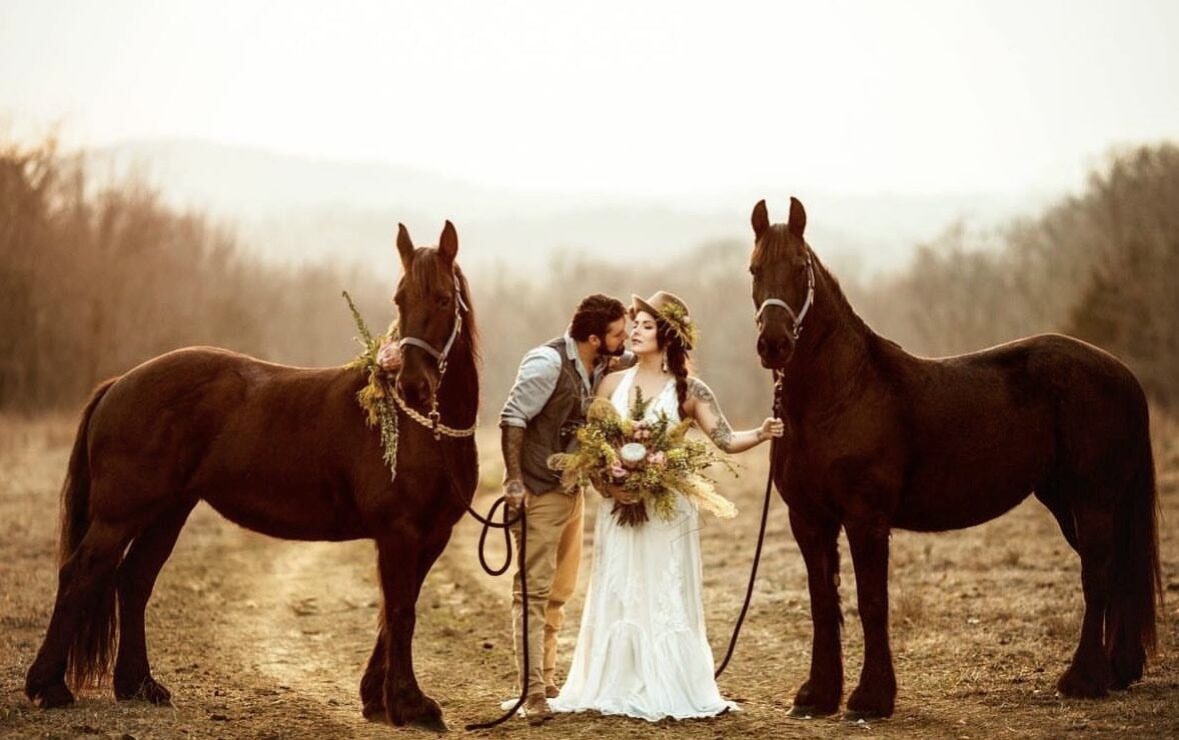 Bride and groom standing between two dark horses in an open field while holding floral arrangements.
