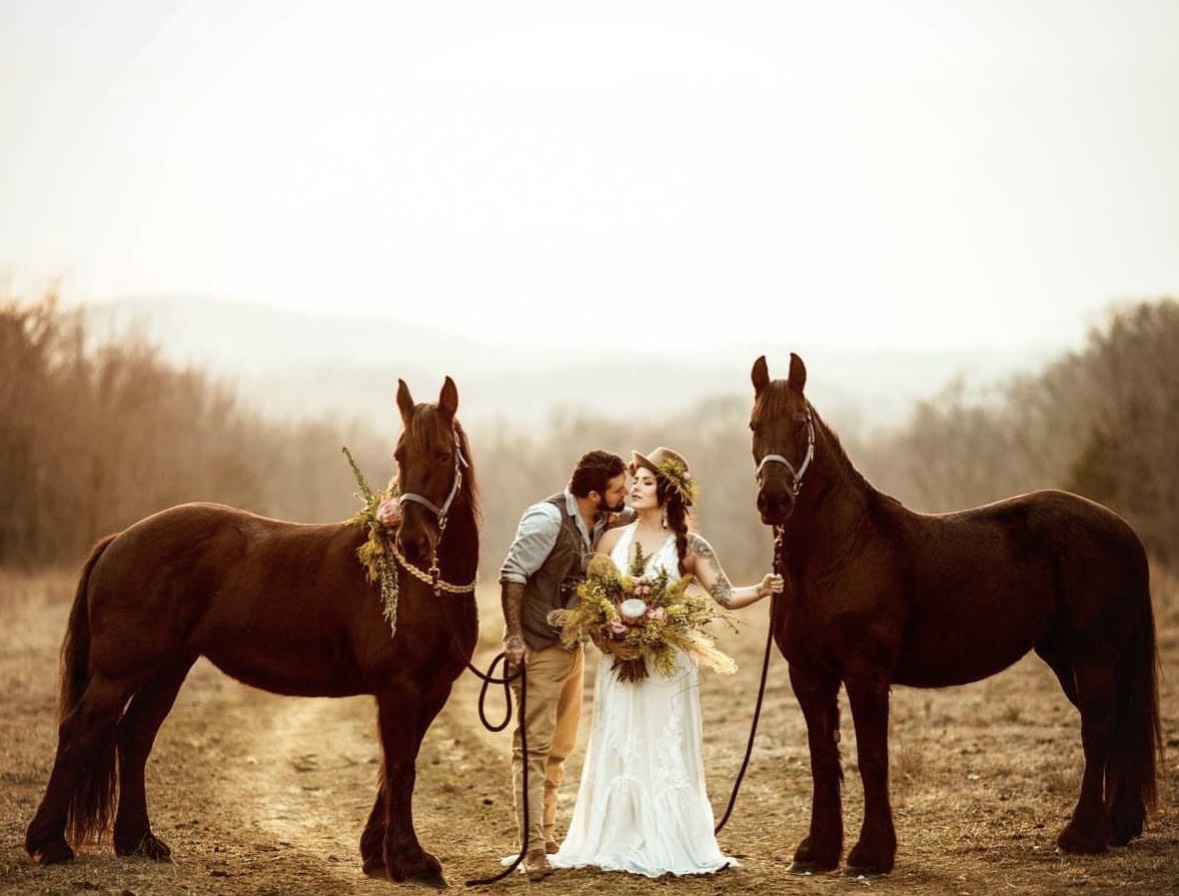 Bride and groom standing between two dark horses in an open field while holding floral arrangements.