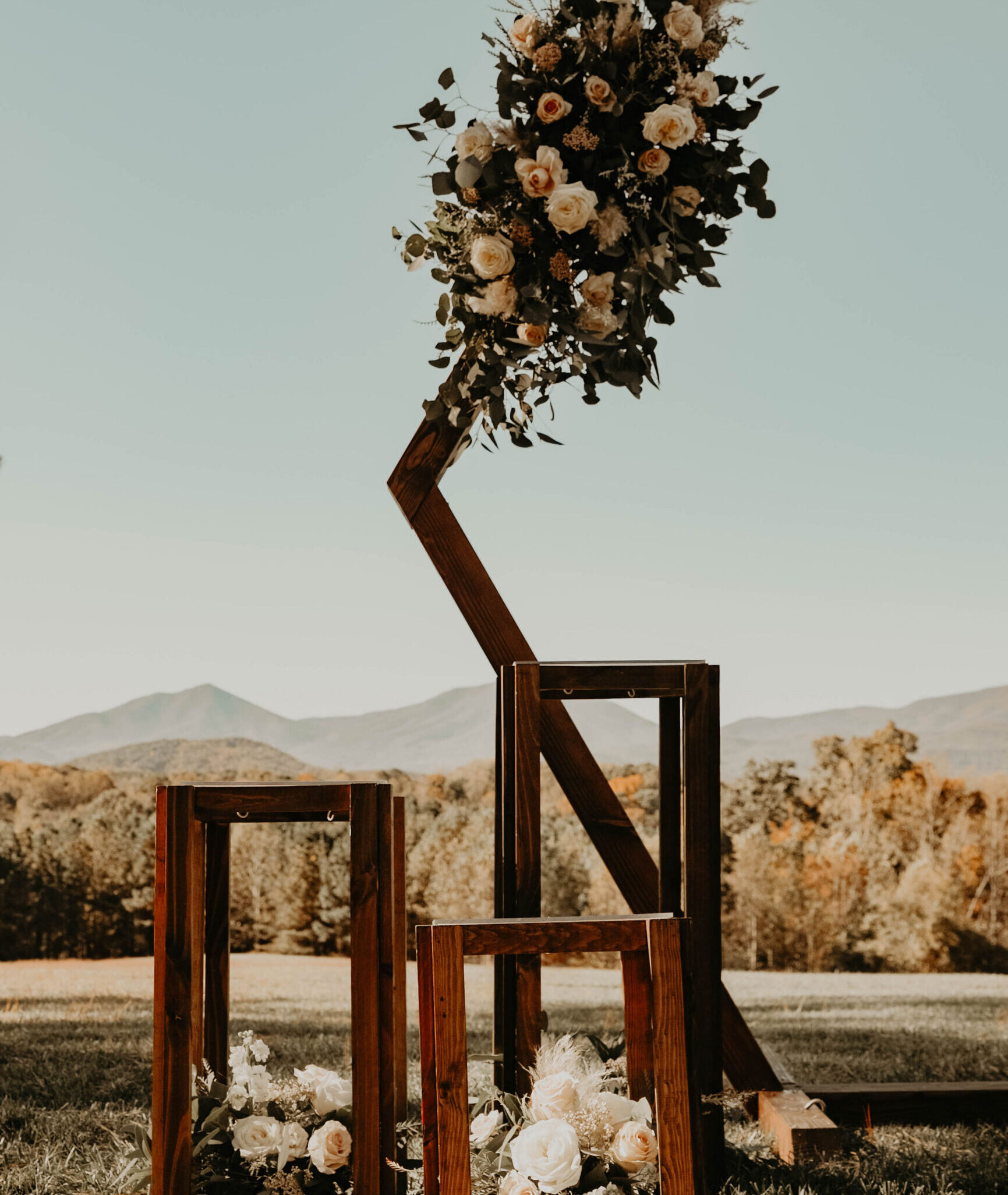 Wooden geometric wedding arch decorated with white flowers and greenery set against a mountain landscape.