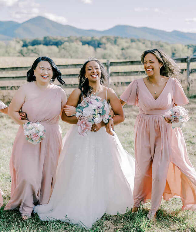 Bride walking arm in arm with bridesmaids in blush pink dresses across a grassy field with mountains behind them.