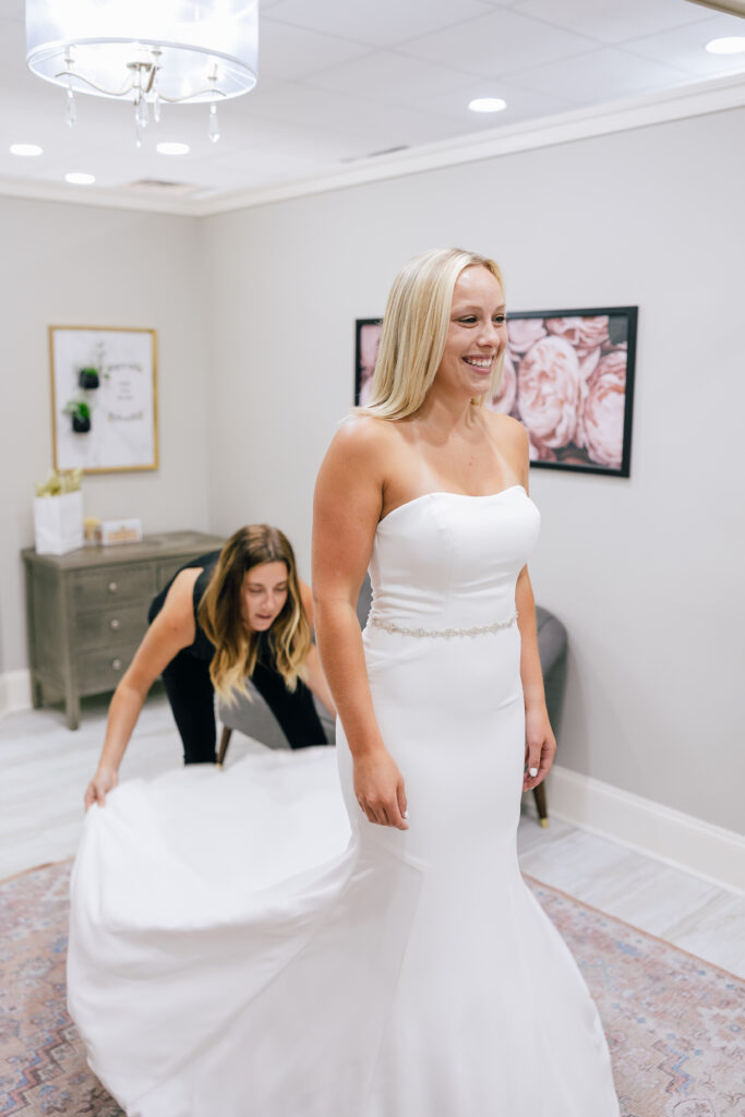 Bride in a strapless fitted wedding gown smiles while a stylist adjusts the train behind her in a fitting room.