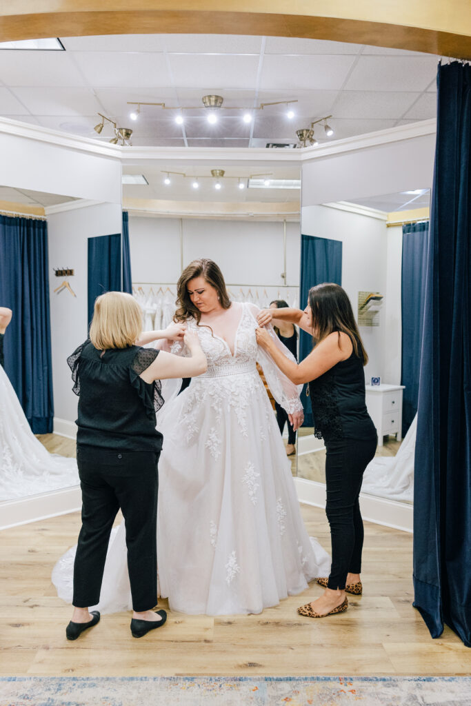 Two stylists adjust the bodice and sleeves of a bride wearing a lace ball gown inside a mirrored bridal suite.